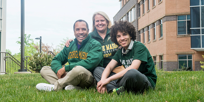 Family of three sitting in a grassy area on campus wearing green Mason shirts